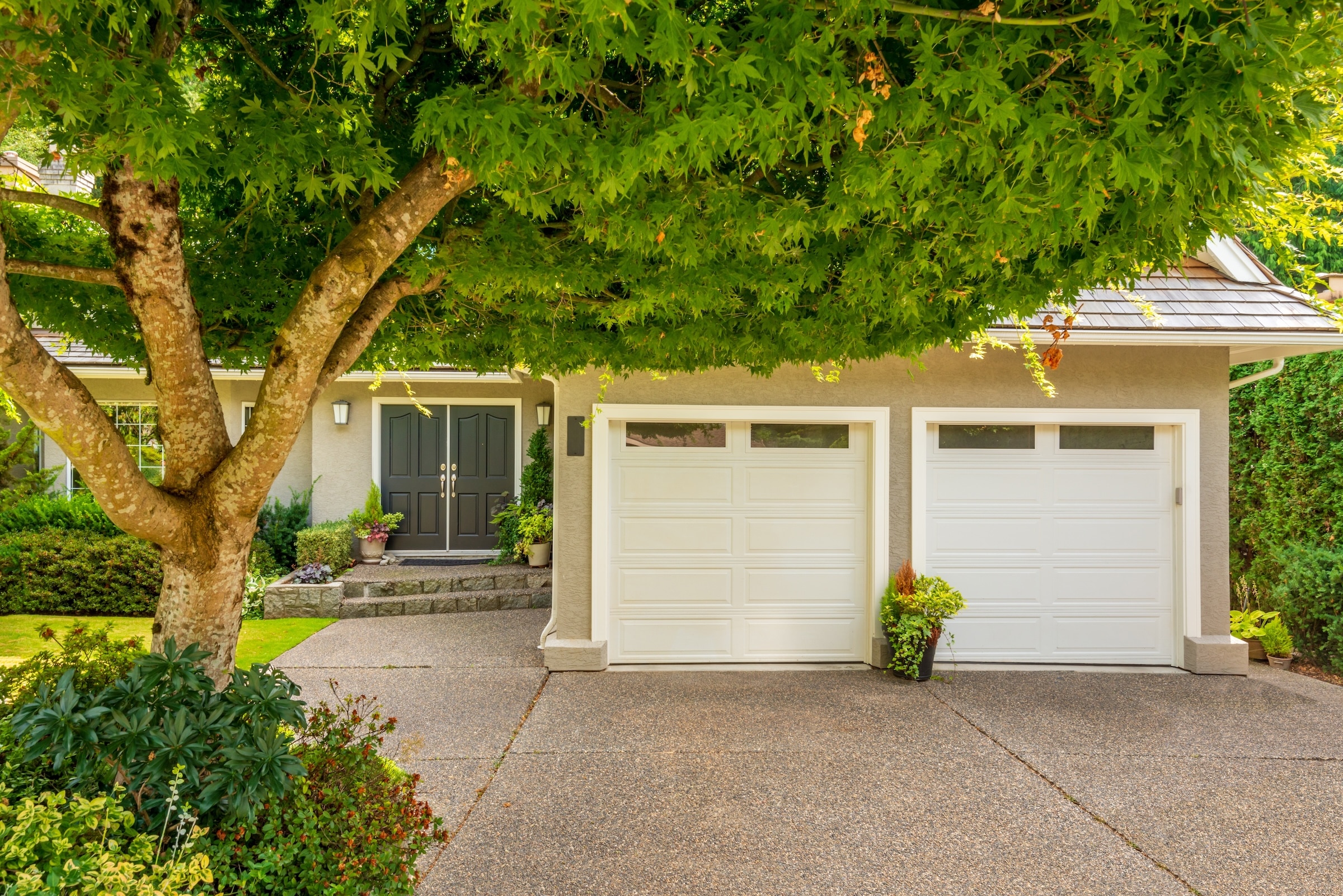 Exterior of house with nice white garage doors and green trees and plants growing in the yard.